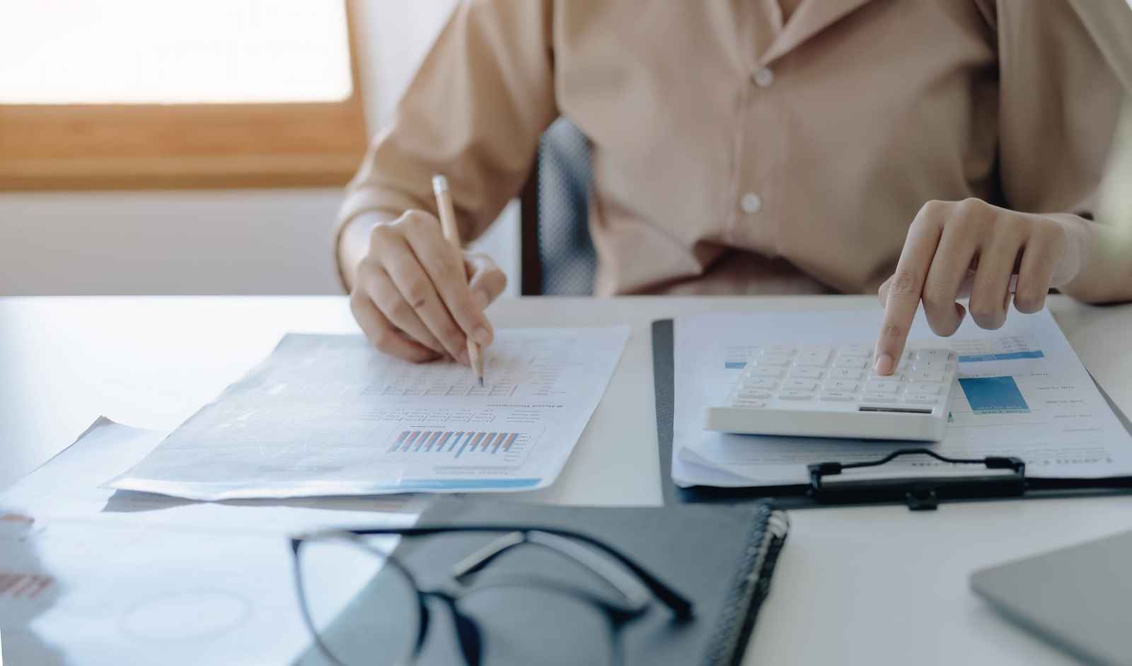 Manos de una persona que anota en un documento con gráficos y usa una calculadora sobre un escritorio