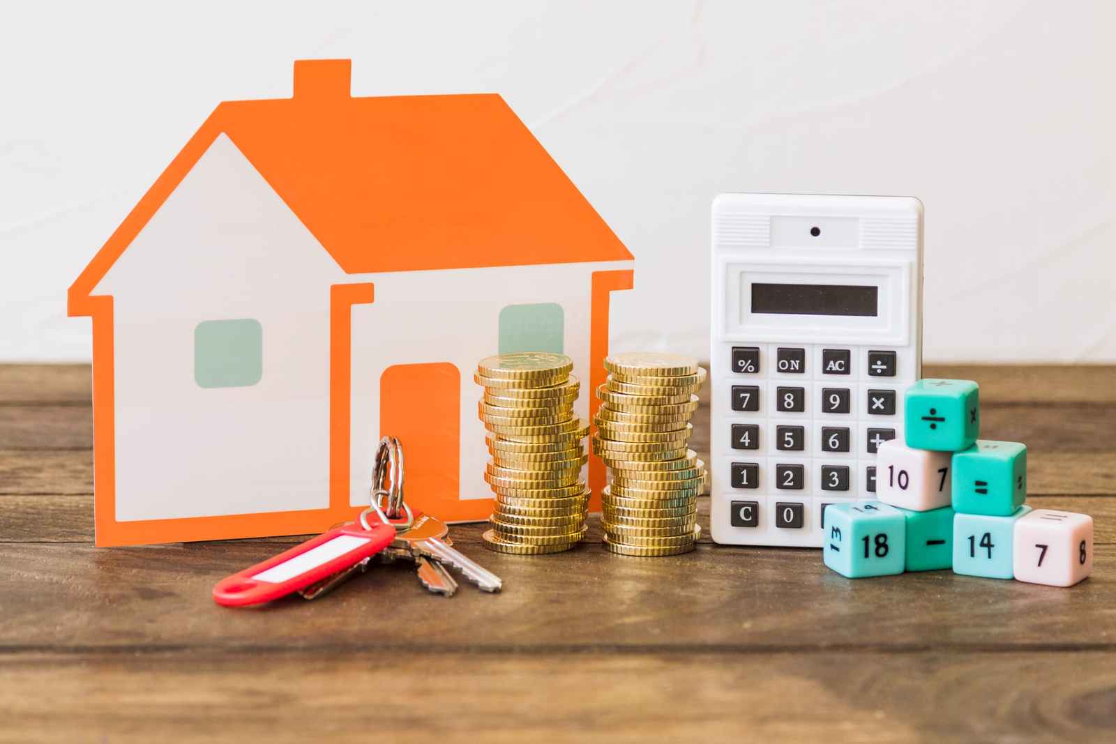 Casa de papel naranja, llaves, monedas apiladas, una calculadora y dados con números sobre una mesa de madera