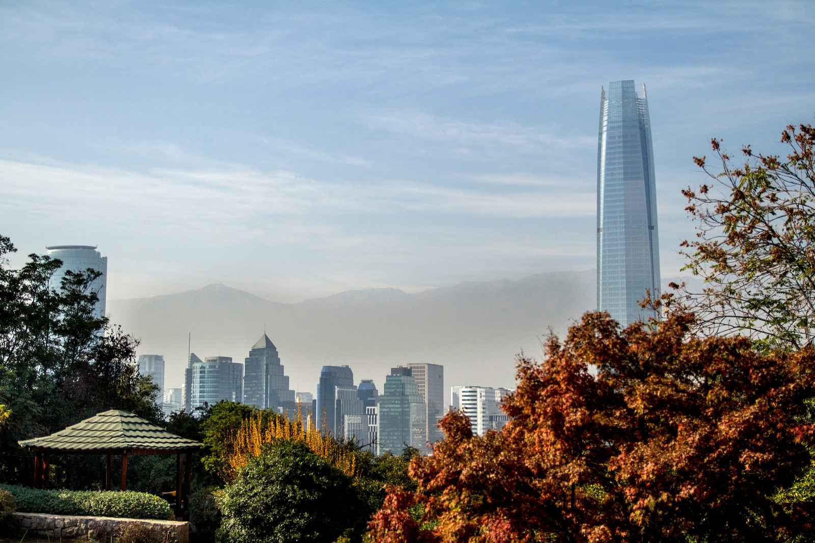 Rascacielos de vidrio y edificios de oficinas de Santiago con la cordillera al fondo, vistos entre árboles de otoño