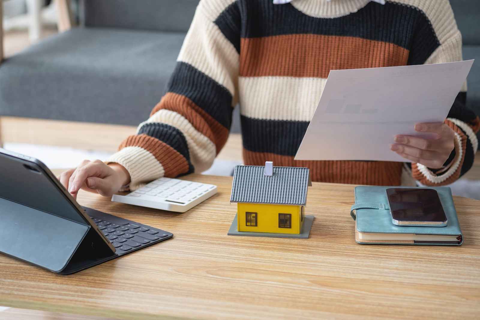 Persona con suéter de rayas revisa un documento y usa una calculadora, con una maqueta de casa amarilla sobre la mesa